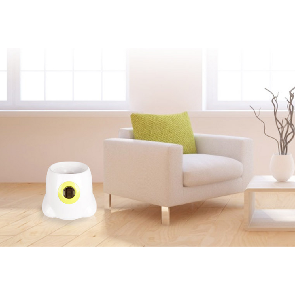 White plastic automatic ball launcher with a yellow opening on a light wood floor next to a white armchair.