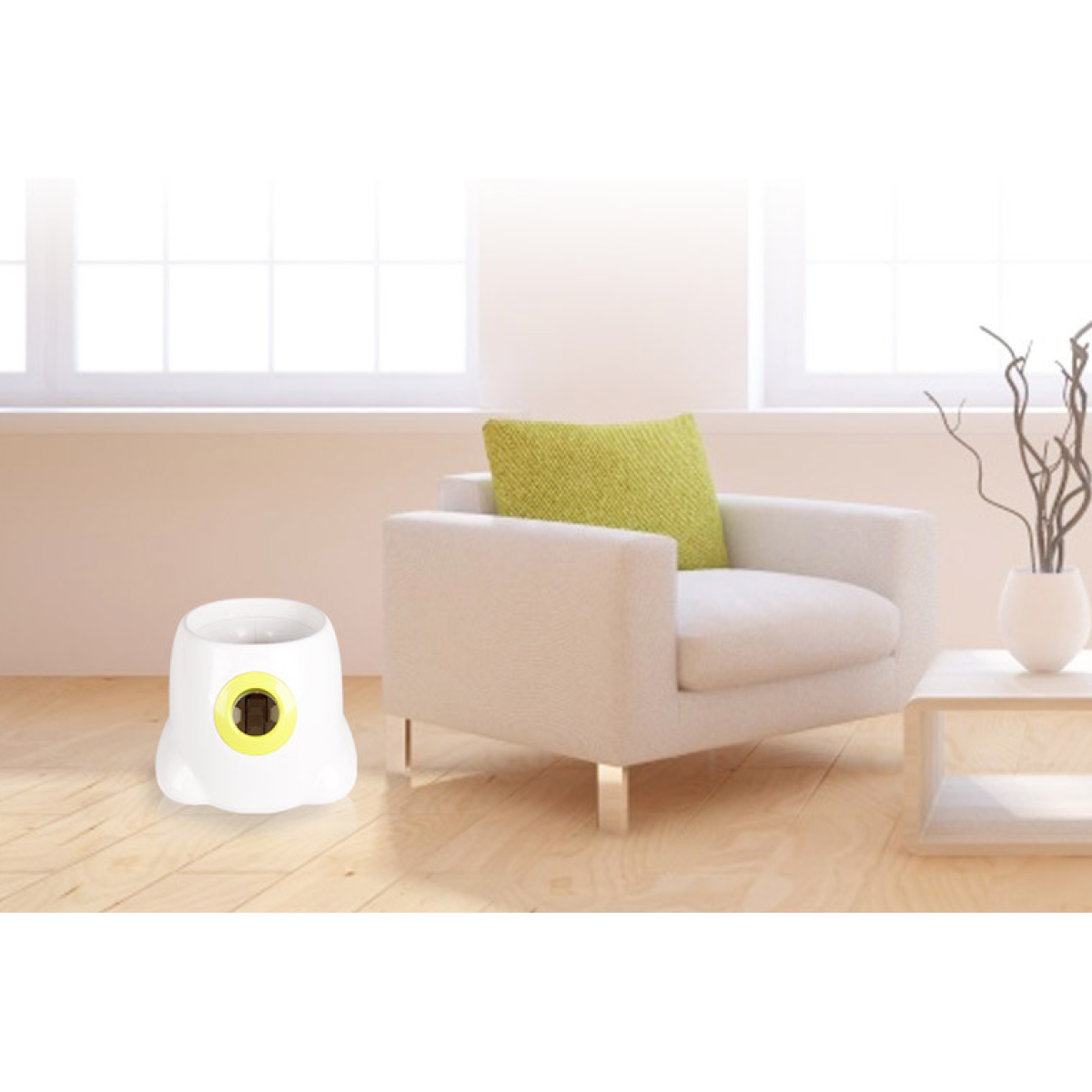 White plastic automatic ball launcher with a yellow opening on a light wood floor next to a white armchair.