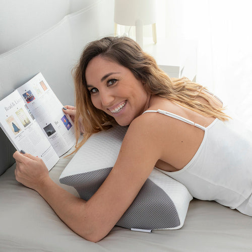 Woman lying on a bed reading a book, resting her arms on a white and grey contoured foam pillow.